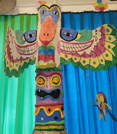 Bunter Totempfahl mit stilisierten Tieren und Gesichtern vor grün-blauem Hintergrund.