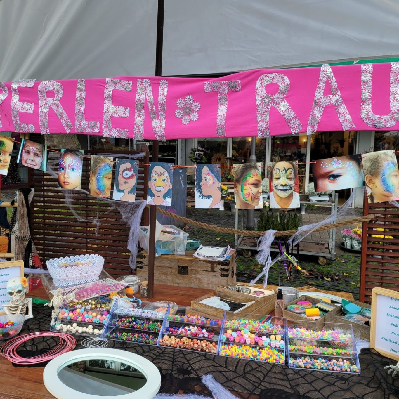 Bunter Marktstand mit Perlenschmuck und Fotos von Gesichtern, hinter einem pinken Banner.