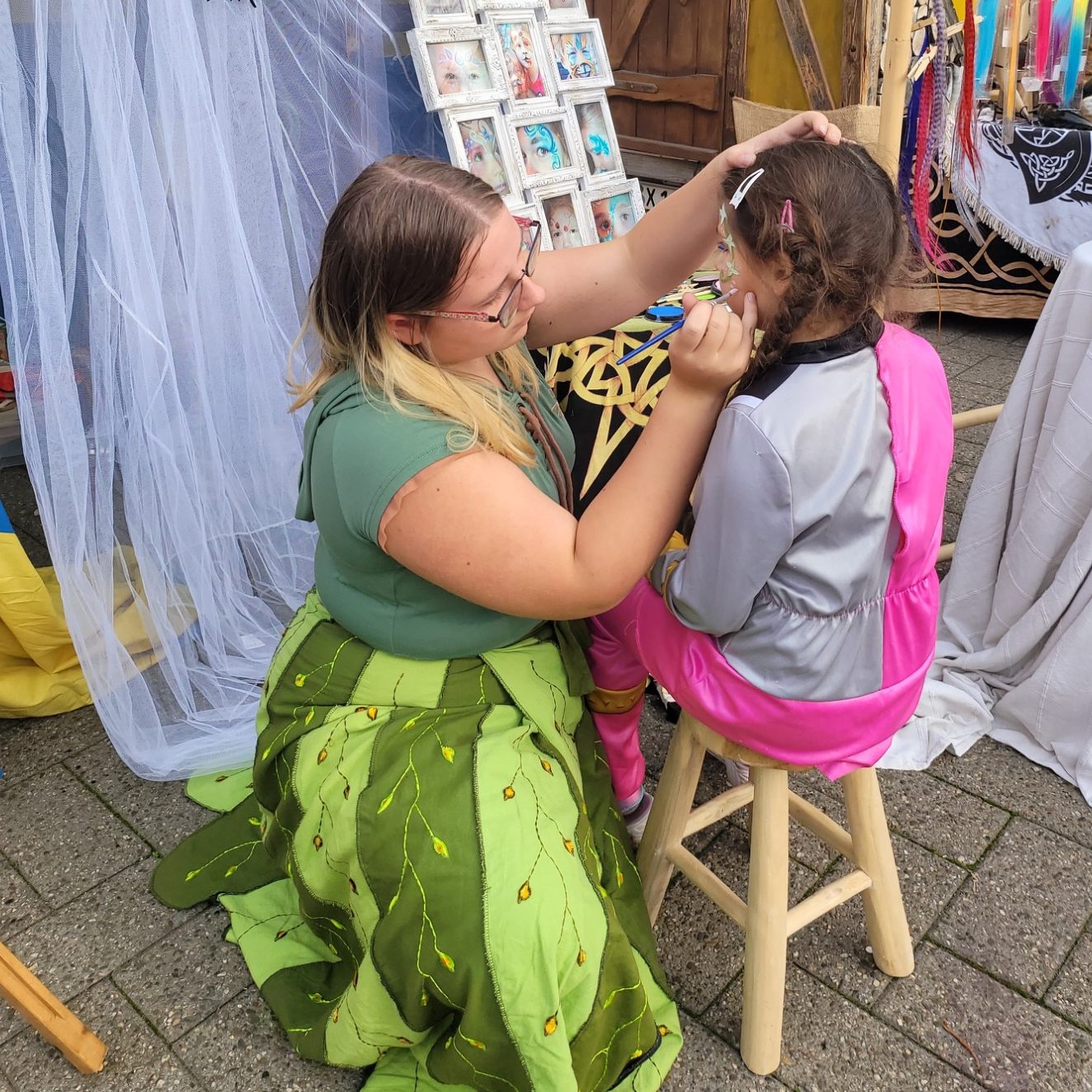 Frau schminkt ein Kind mit einem farbenfrohen Kostüm auf einem Hocker.