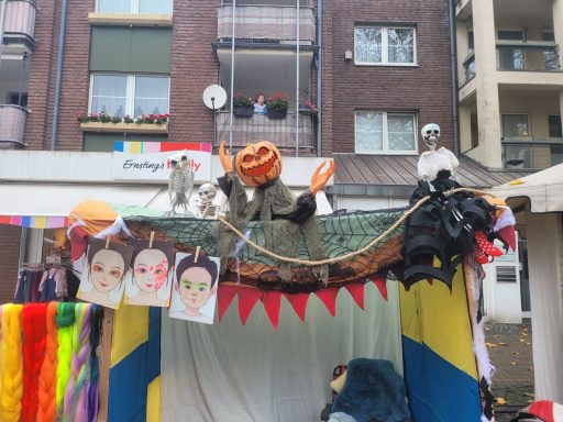 Bunte Festdekoration mit Kürbis, Masken und gemalten Gesichtern vor einem Gebäudebalkon.