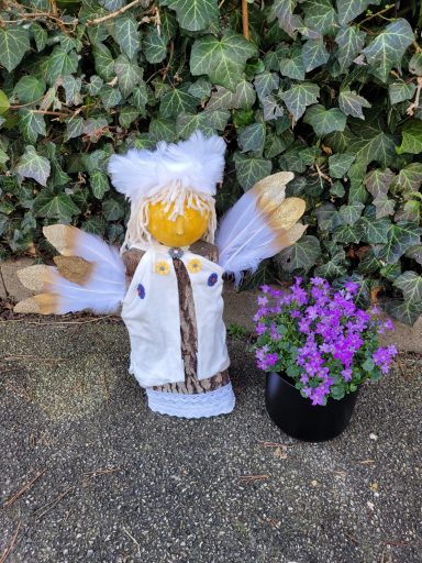 Eine bunte Puppe mit Flügeln steht neben einem Topf mit lila Blumen.