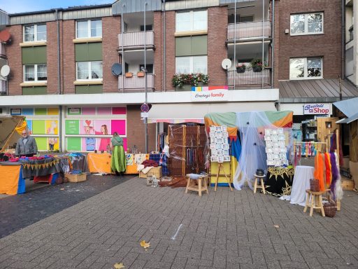 Markt mit bunten Ständen, Textilien und Gebäuden im Hintergrund.