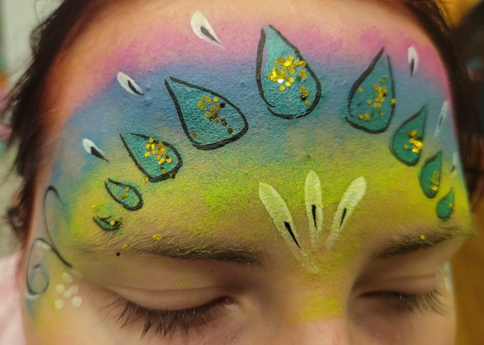 Bunte Gesichtsbemalung mit Tropfen- und Blütenmotiven auf der Stirn.