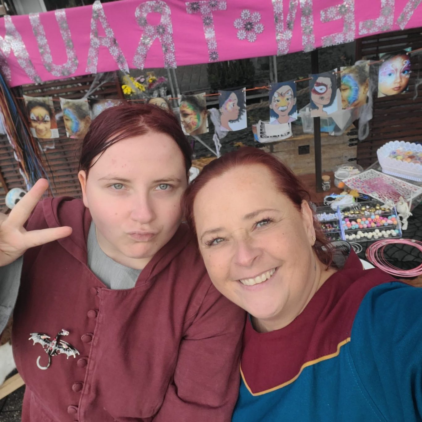 Zwei Frauen in mittelalterlicher Kleidung posieren vor einem Stand mit bunten Kunstwerken.