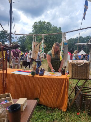 Eine Frau arbeitet an einem Holztisch auf einem Markt mit bunten Ständen im Hintergrund.