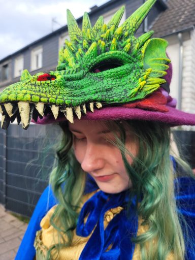 Person mit grüner Drachenmütze und lockigen Haaren, trägt ein blaues und goldenes Outfit.