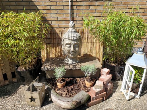 Buddha-Kopfskulptur umgeben von Pflanzen und dekorativen Elementen in einem Garten.