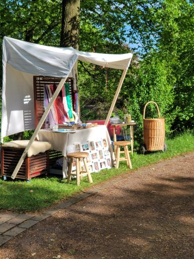 Ein Verkaufsstand mit Tisch, Stühlen und bunten Stoffen, umgeben von Bäumen.