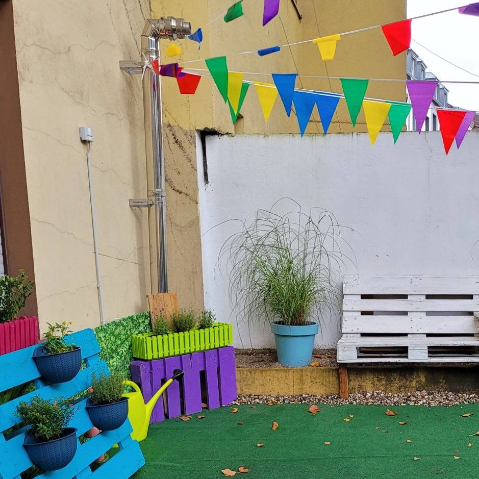 Bunte Girlanden hängen über einem bunten Garten mit Pflanzen und einer Sitzbank.