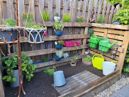 Kleiner Gartenbereich mit Blumenkästen, Pflanzen und Zubehör wie Gießkanistern und Eimern.