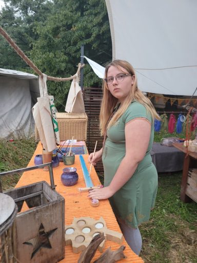 Junge Frau in einem grünen Kleid steht an einem Tisch mit Farben und Handwerksmaterialien.