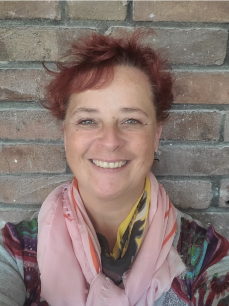 Frau mit rotem Haar, lächelt, trägt ein buntes Tuch und steht vor einer Steinmauer.