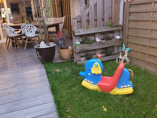 Bunte Wippe in einem Garten mit Holzdecks und blühenden Pflanzen im Hintergrund.