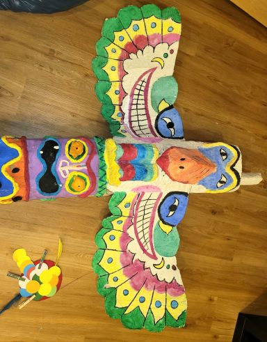 Bunt bemalter Vogel mit großen Augen und farbenfrohen Mustern auf Holzplatte.