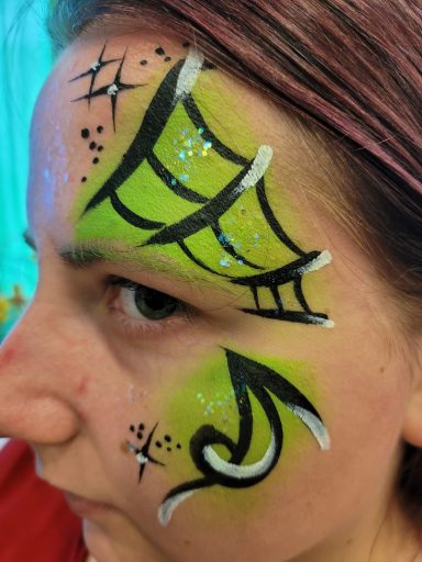 Gesichtsbemalung mit grünem Spinnennetz, schwarzem Pfeil und Glitzerakzenten.